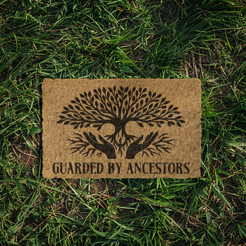 18x24 inch rectangular coir doormat made from natural coconut husk fibers, featuring a bold black print of a stylized tree with branching leaves and intertwined roots forming the shape of protective hands. Below the tree design, the phrase "GUARDED BY ANCESTORS" is printed in large, capitalized serif lettering. The mat has a textured, bristled surface ideal for scraping dirt and moisture from shoes, and a durable construction suited for everyday entryway use. Designed for indoor or outdoor placement, this d